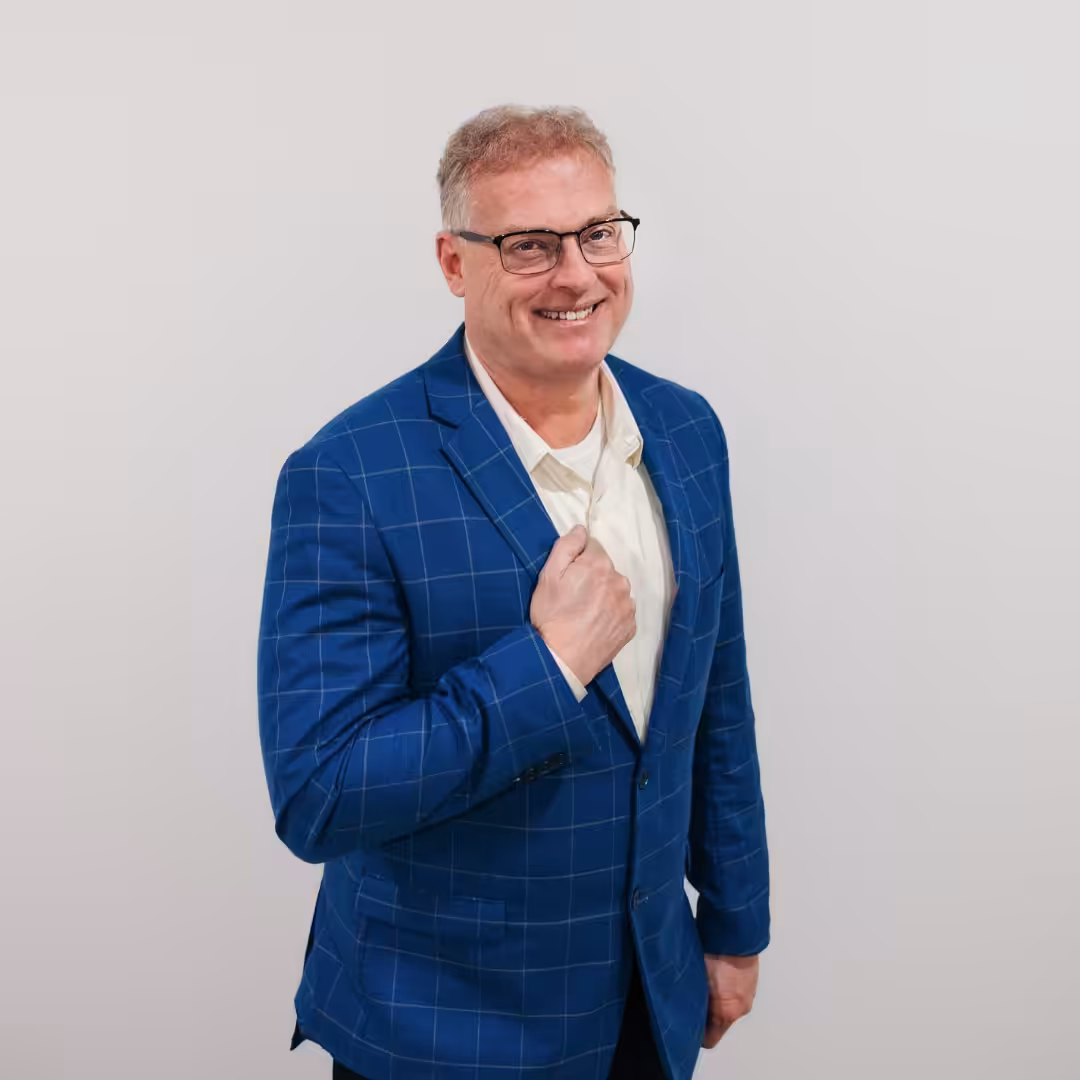 A smiling man with short gray hair and glasses wearing a blue checkered suit jacket over a white shirt poses against a plain white background.