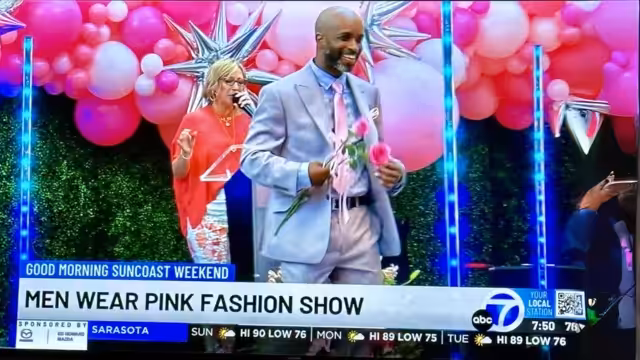 A man in a light pink suit holds pink flowers and smiles while walking on stage at a fashion show. A woman in a red outfit stands behind him, speaking into a microphone. Pink balloons and star decorations fill the background.