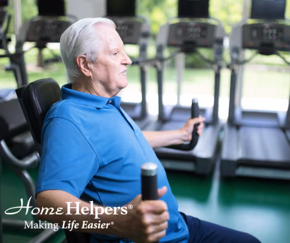 An older man with white hair uses a chest press machine at a gym. He wears a blue polo shirt and sits in front of treadmills. Home Helpers Making Life Easier is written in the lower left corner.