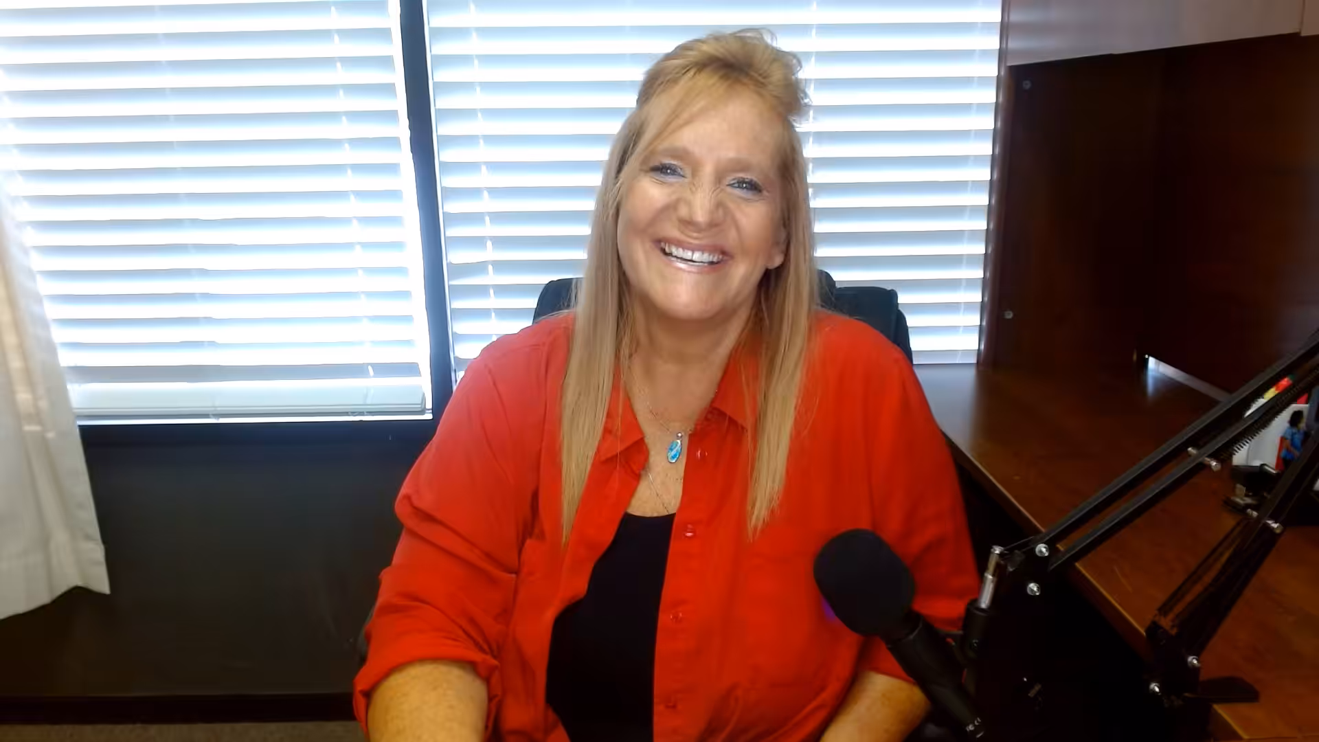 A woman with long blonde hair, wearing a red shirt over a black top, sits at a desk smiling at the camera. A microphone is positioned on the desk next to her. Blinds cover the window behind her.