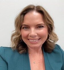 A woman with wavy, shoulder-length light brown hair is smiling at the camera. She is wearing a teal blazer, hoop earrings, and a delicate necklace, with a plain light background.