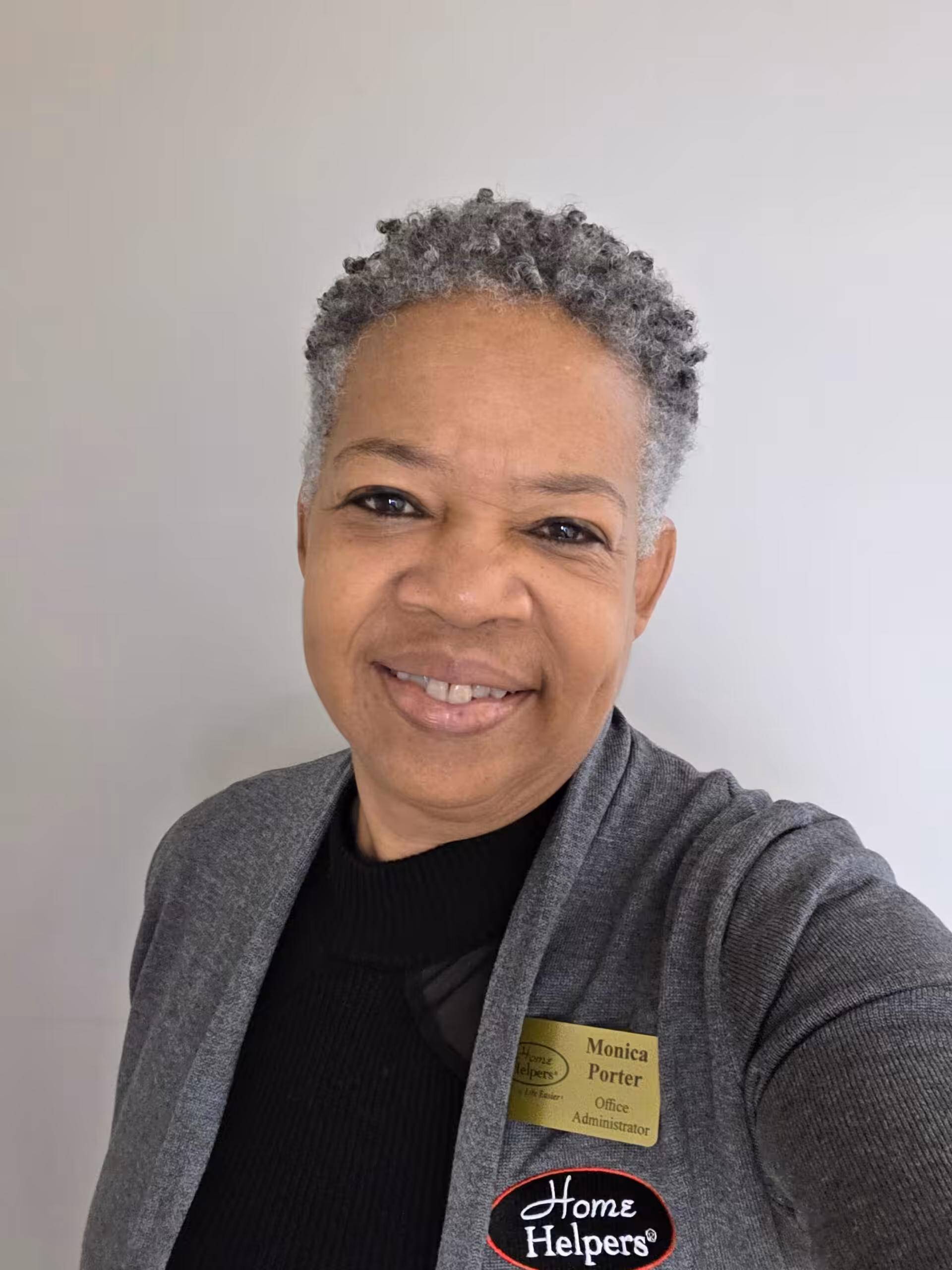 A smiling woman with short gray hair wearing a gray cardigan, black top, and a name badge that reads Monica Porter, Office Administrator. A Home Helpers logo is visible on her outfit. She is standing in front of a plain, light background.