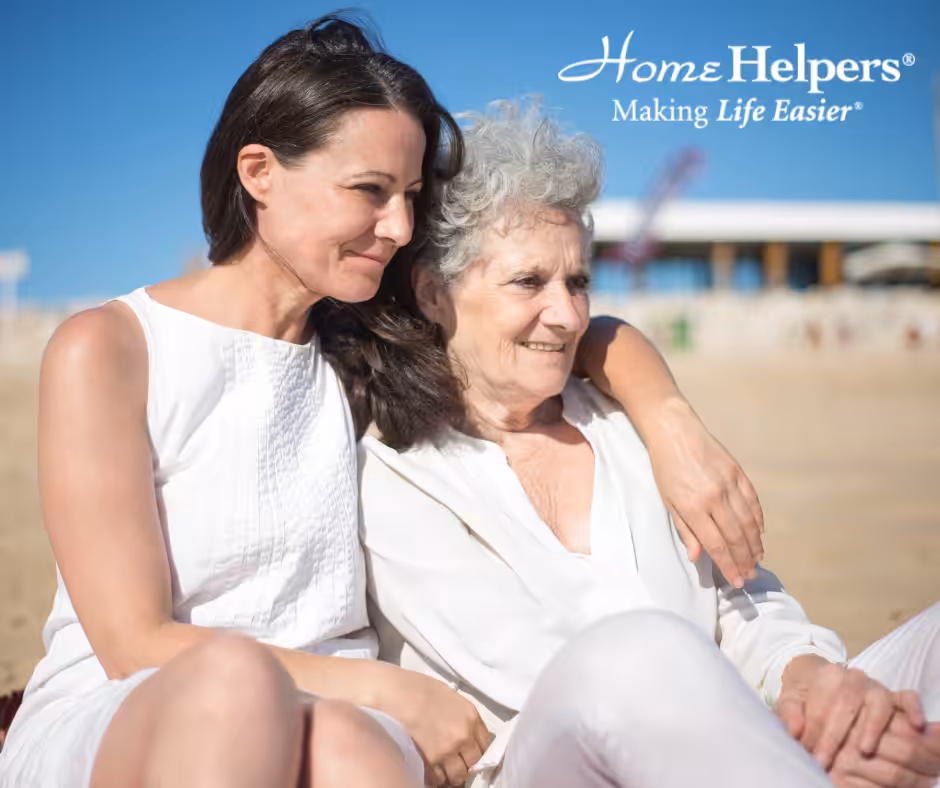A younger woman hugs an older woman as they sit together outdoors on a sunny day, both smiling and wearing white. The text Home Helpers® Making Life Easier® is displayed in the top right corner.