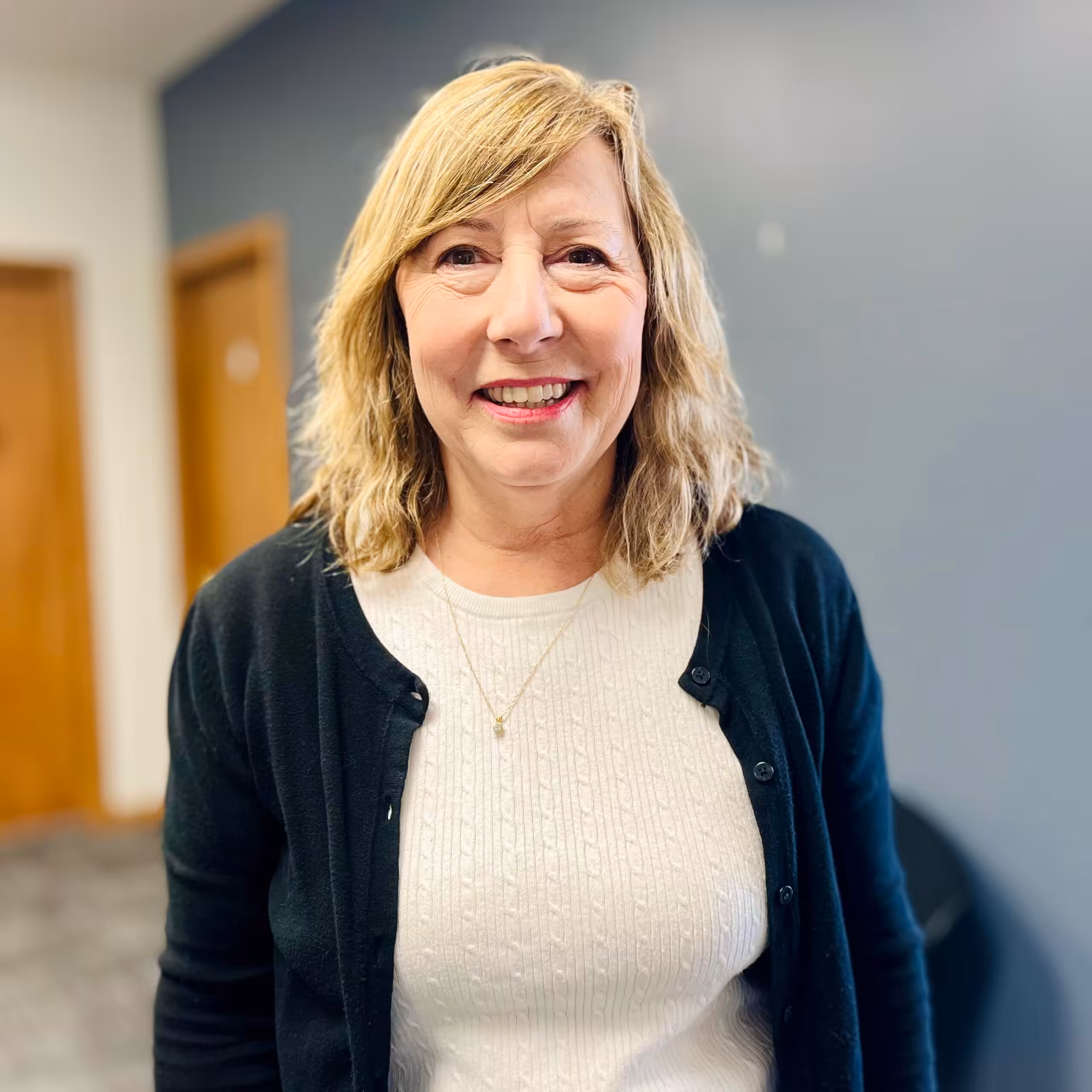 A woman with blonde, shoulder-length hair smiles at the camera. She is wearing a cream-colored sweater and a black cardigan, standing indoors against a gray and white wall.