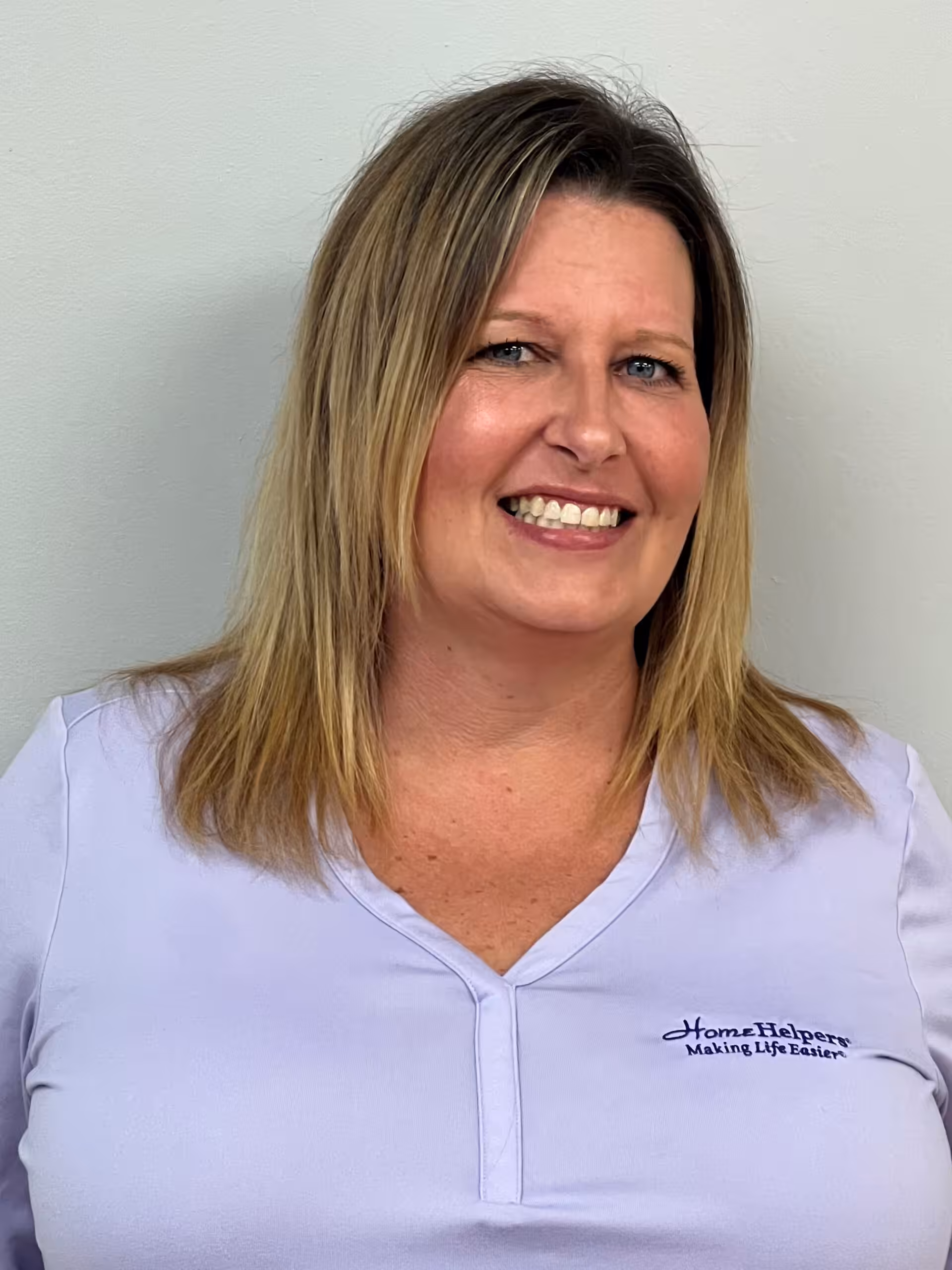 A woman with blonde hair smiles at the camera. She is wearing a light purple shirt with a Home Helpers Making Life Easier logo on the left side. The background is plain and light-colored.