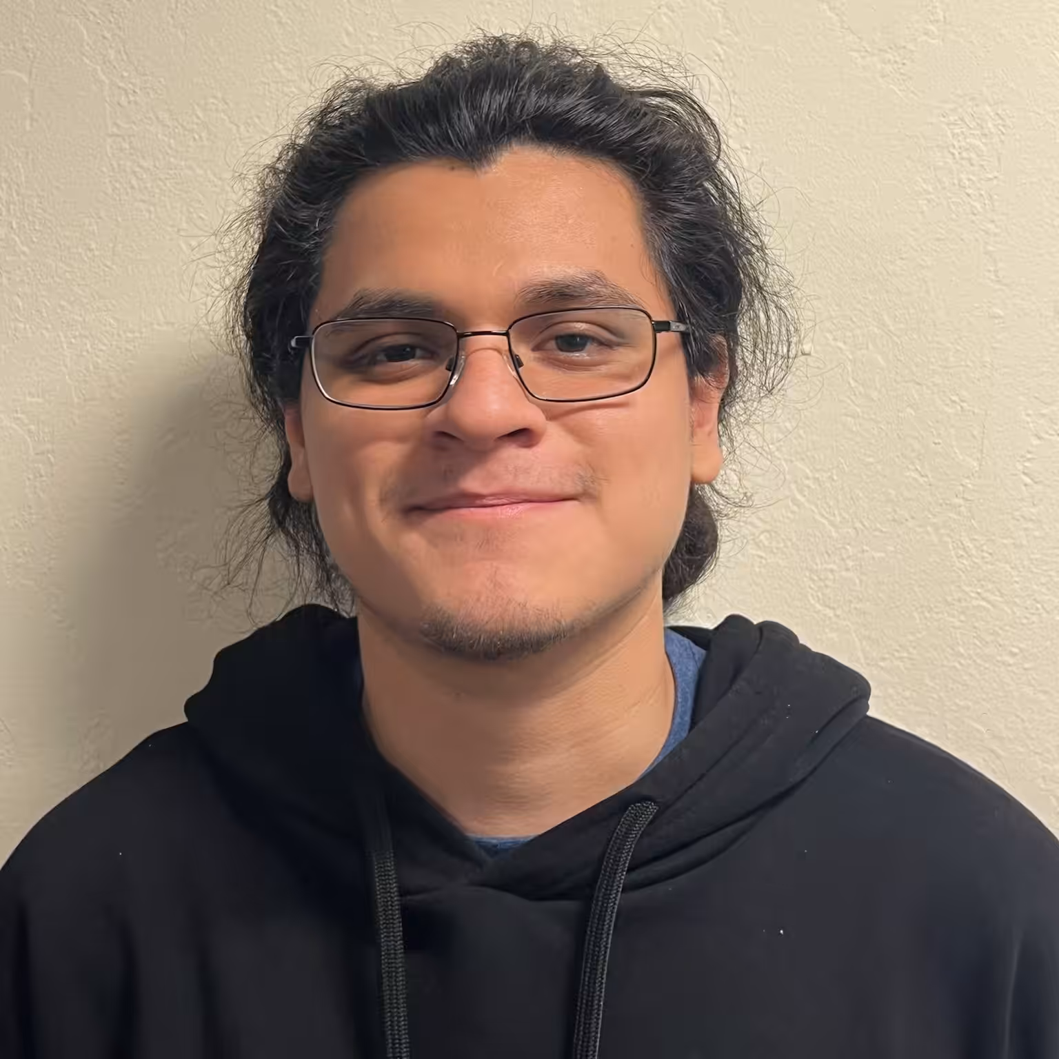 A person with long dark hair, glasses, and slight facial hair is smiling while wearing a black hoodie, standing in front of a textured beige wall.