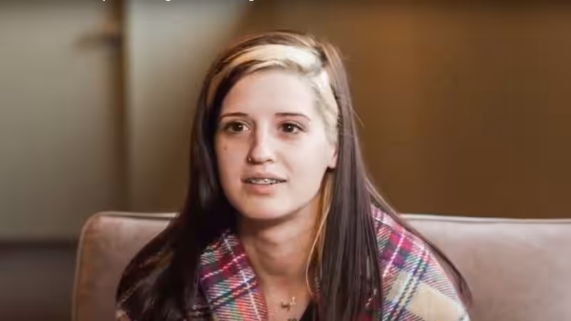 A young woman with long brown hair and a streak of blonde sits on a beige couch, wearing a colorful plaid jacket. She is looking slightly off-camera and smiling softly. The background is out of focus.