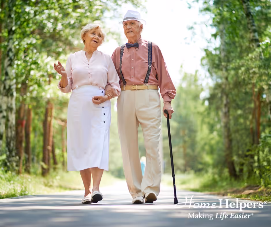 An elderly couple walks arm in arm down a sunny, tree-lined path. The woman smiles and talks while the man, wearing suspenders and a hat, uses a cane. Both appear happy and relaxed. The image includes the Home Helpers logo.