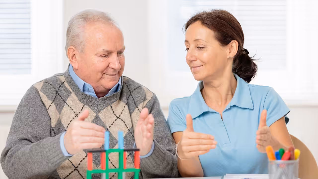 An older man and a woman sit at a table playing a game with colorful rods and blocks. They smile and gesture with their hands, appearing engaged and cheerful in a bright, sunlit room.