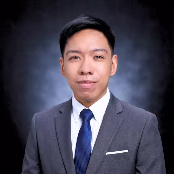 A young man in a gray suit, white shirt, and blue tie poses for a formal portrait against a dark, gradient background. He has short black hair and is smiling slightly.