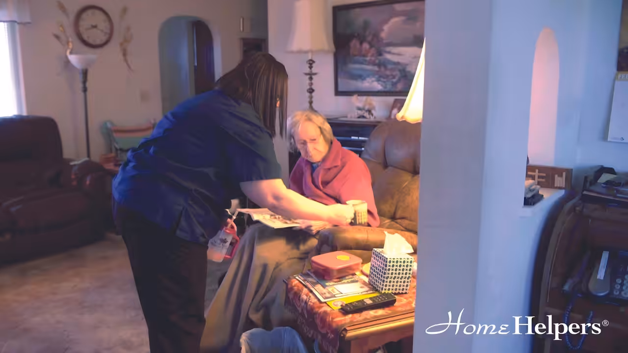 A caregiver hands a drink to an elderly woman sitting in a recliner in a cozy living room. Books, tissues, and other items are on the side table. The Home Helpers® logo is visible in the lower right corner.