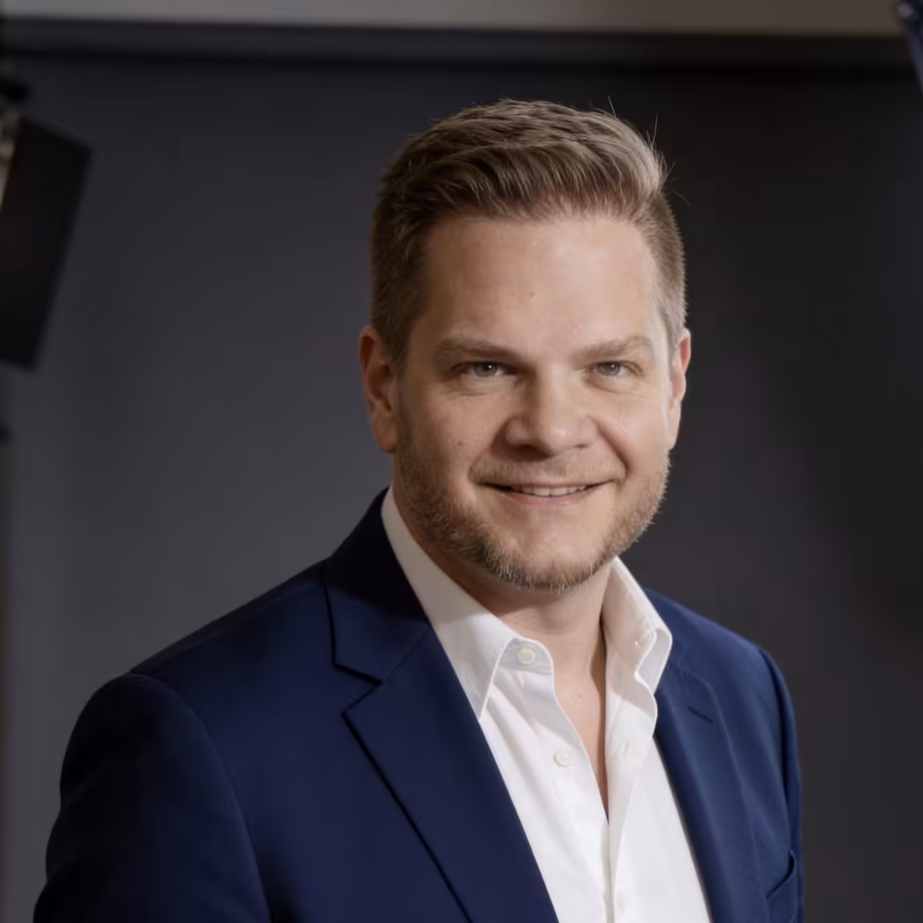 A man with short light brown hair and a trimmed beard smiles at the camera. He is wearing a dark blue suit jacket over a white shirt, standing against a dark, studio-style background.