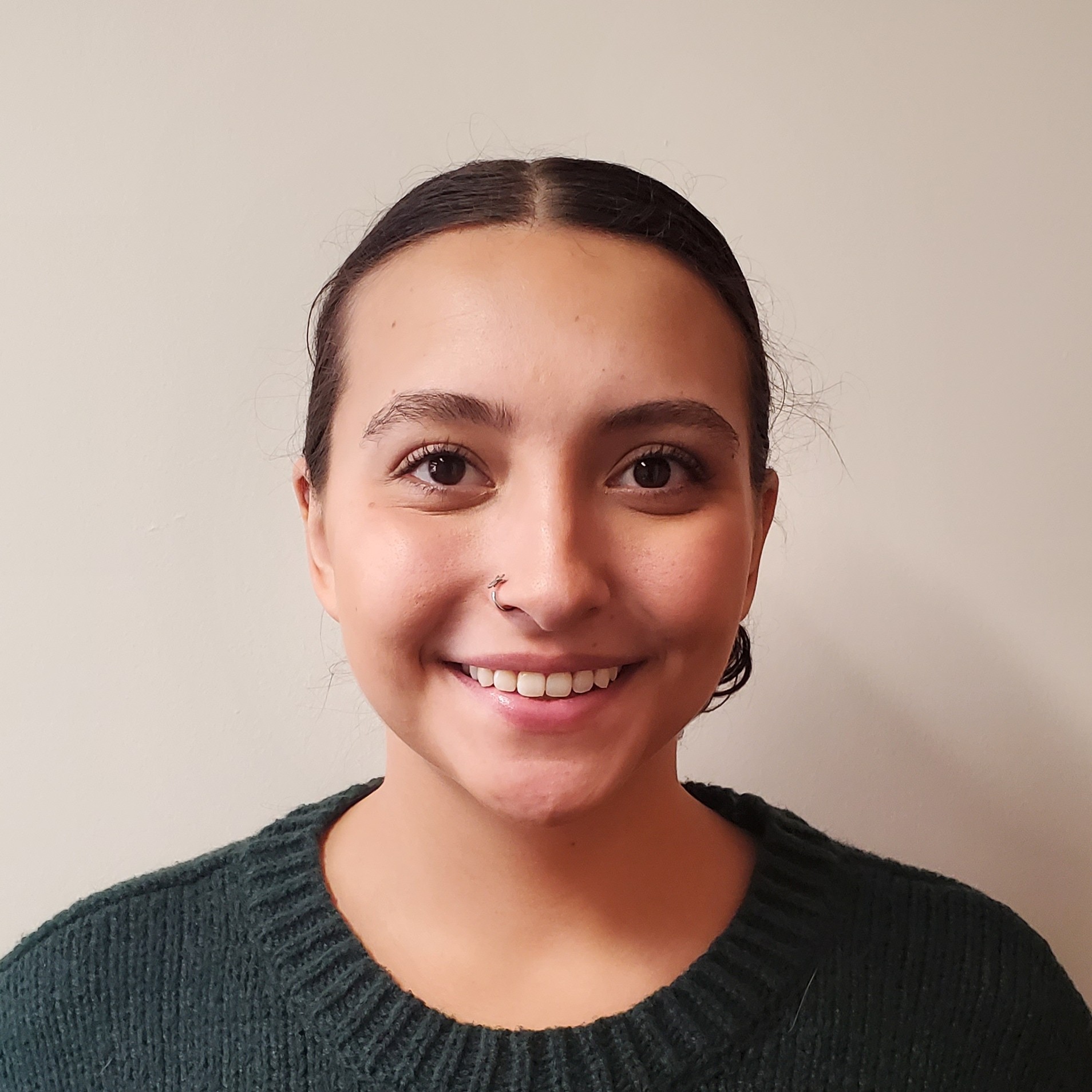 A young woman with dark hair pulled back, wearing a green sweater and a nose ring, smiles at the camera in front of a plain light-colored background.