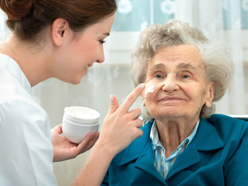 A young caregiver smiles while applying cream to the face of an elderly woman with gray hair, who is wearing a blue jacket and smiling gently.
