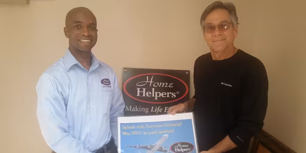 Two men stand indoors, smiling. One holds a poster for a Summer Getaway contest, showing an airplane. Behind them is a Home Helpers® Making Life Easier® sign on the wall.