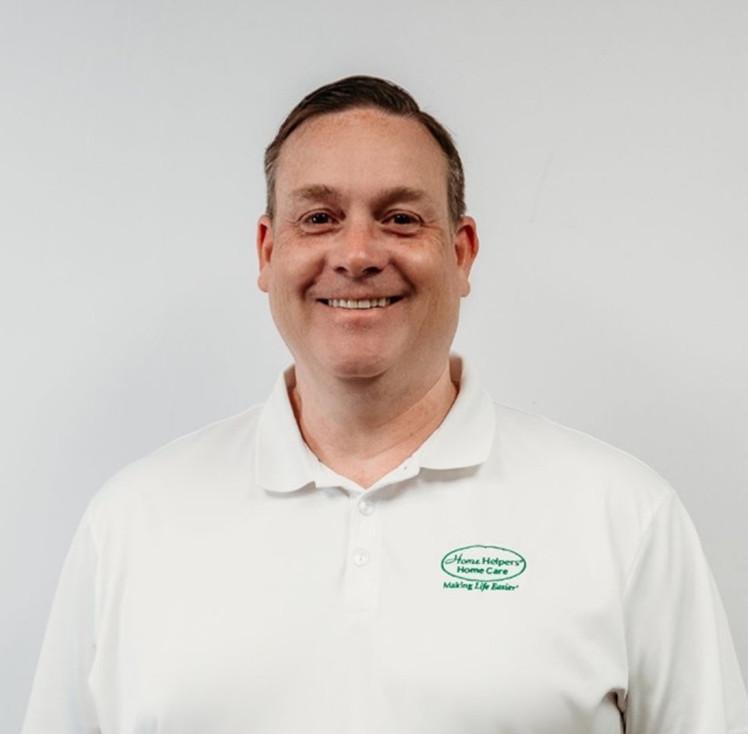 A smiling man with short brown hair wears a white polo shirt with a green Home Helpers Home Care logo, standing in front of a plain white background.