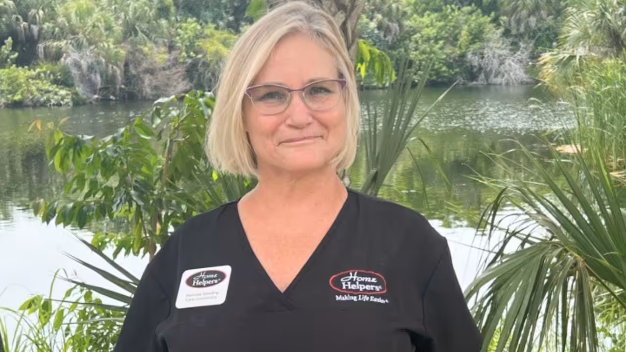 A woman with blonde hair and glasses, wearing black Home Helpers scrubs, stands outdoors in front of a lake surrounded by green trees and plants. She is smiling at the camera.