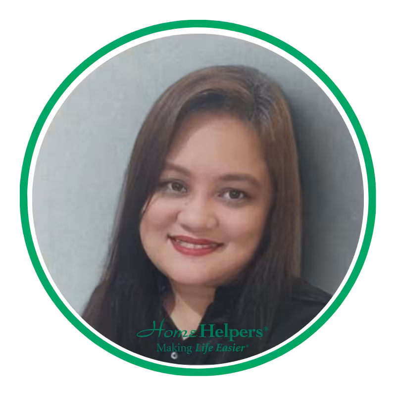 Portrait of a woman with long brown hair, smiling, wearing a black top. The image has a green circular border with the Home Helpers logo and the slogan Making Life Easier at the bottom.