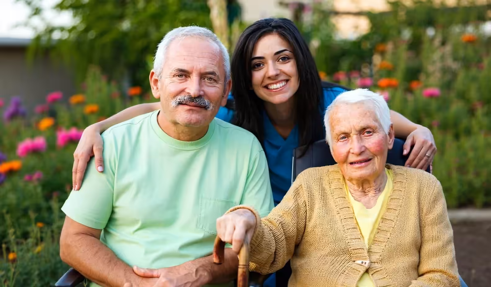 A smiling young woman stands behind an elderly man and woman sitting outdoors in a garden, with flowers in the background. The woman in the middle has her arms around both seniors.