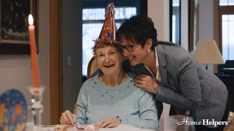 An elderly woman wearing a birthday hat smiles at a table with cake, while another woman in a blazer leans in and smiles beside her. The Home Helpers logo is visible in the corner.