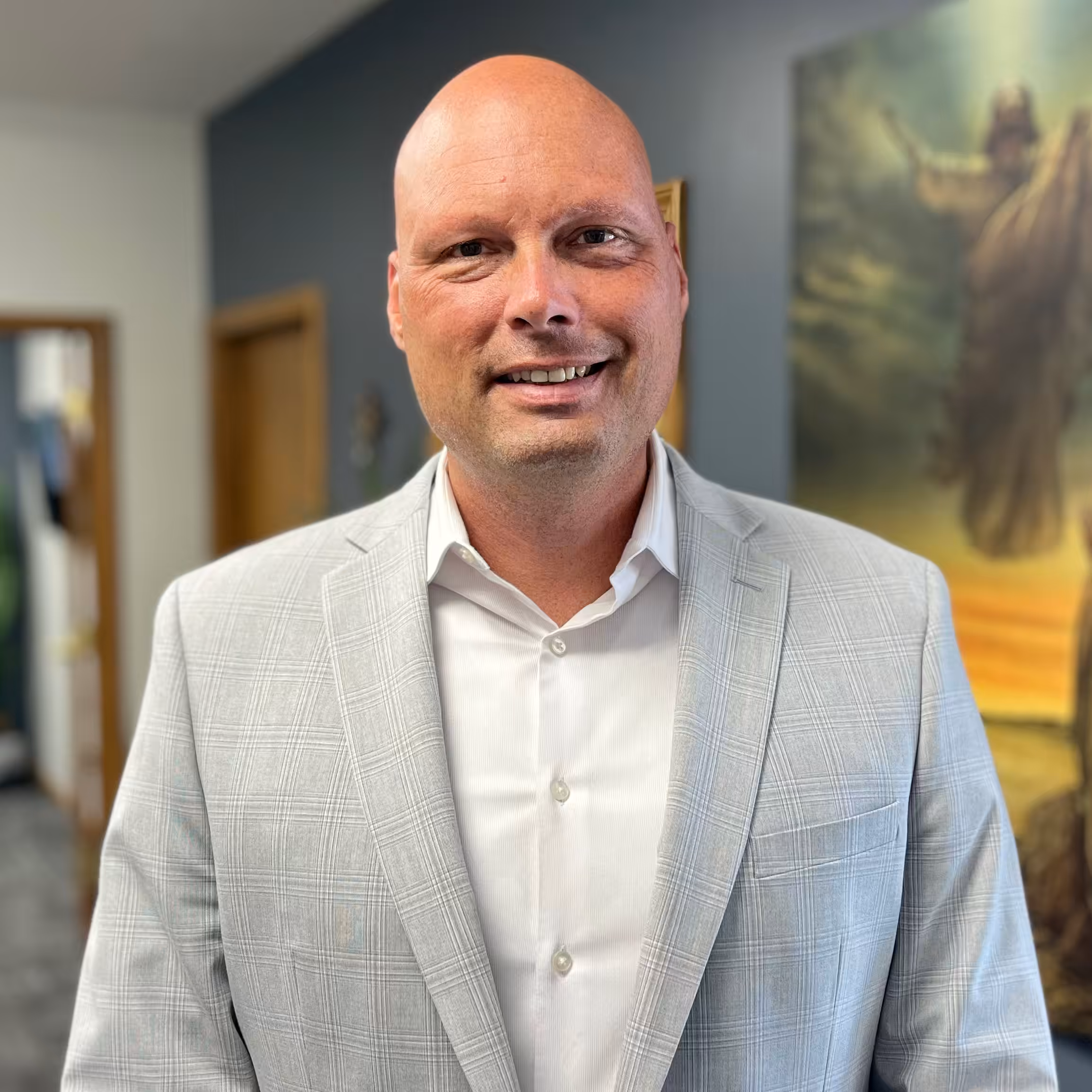 A smiling bald man wearing a light gray plaid suit jacket and white shirt stands indoors, with a blurred painting and doorway visible in the background.