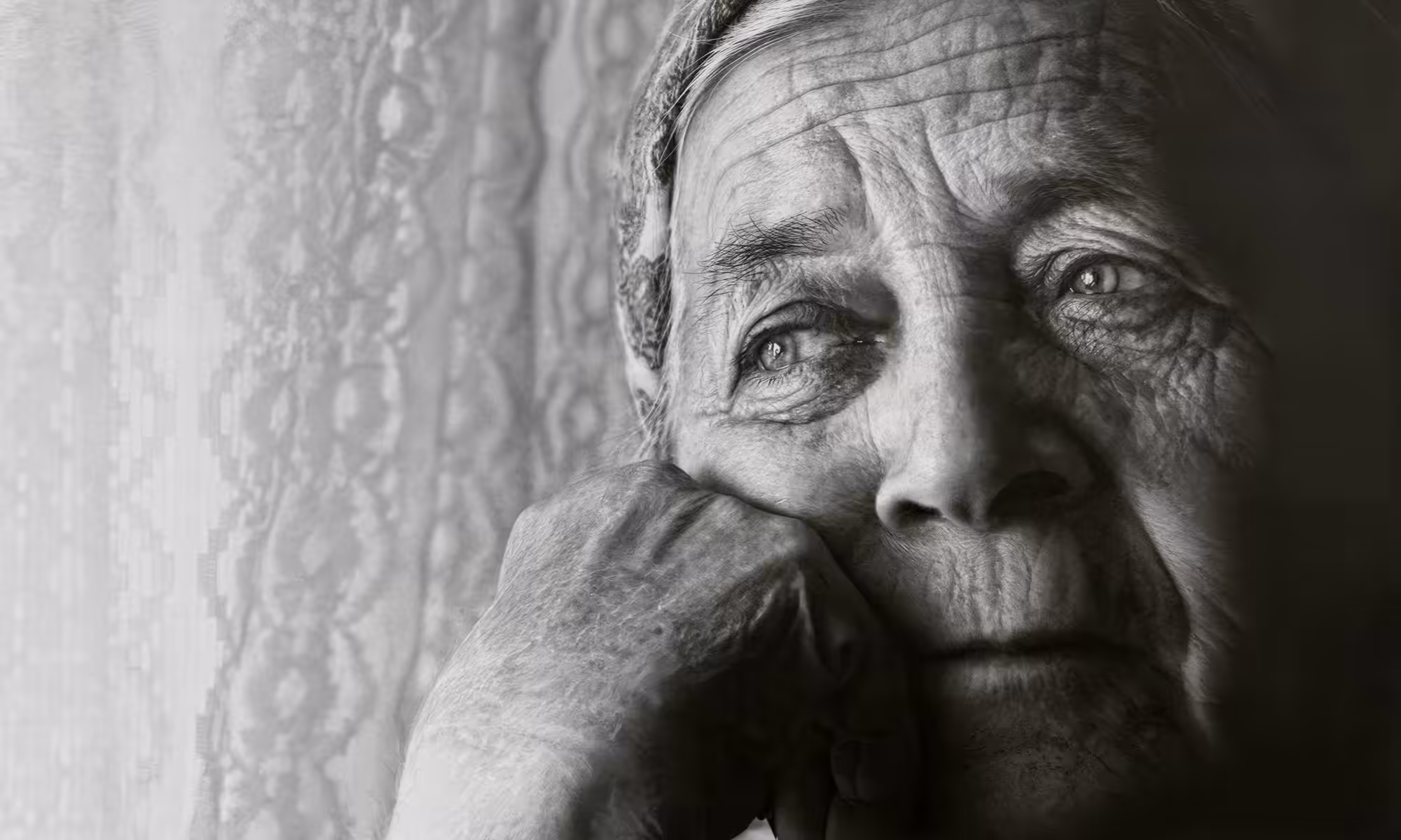 Black and white close-up of an elderly person staring thoughtfully out a window, their hand resting on their chin, highlighting deep wrinkles and an expression of contemplation or nostalgia.