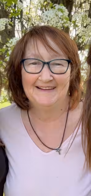 A smiling woman with short brown hair and glasses stands outdoors in front of greenery and white flowers, wearing a light pink top and a necklace.