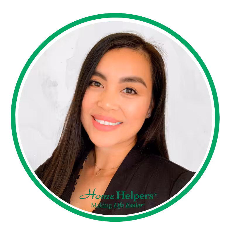 A woman with long dark hair, wearing a black blazer and a necklace, smiles at the camera. The image has a white background with a green circular border and the Home Helpers logo at the bottom.