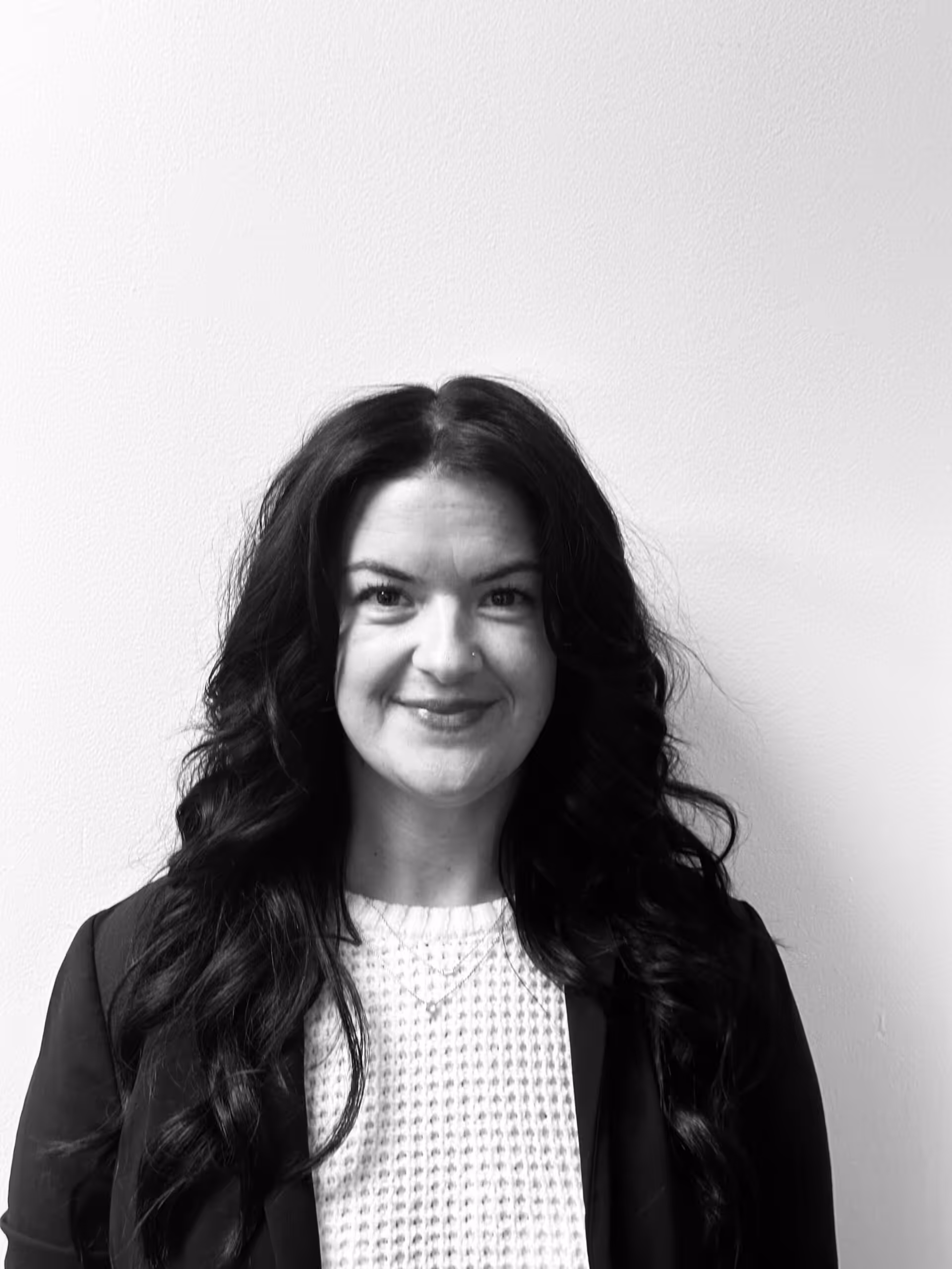 Black and white photo of a woman with long, wavy dark hair, wearing a light textured top and a dark jacket, smiling at the camera against a plain light-colored background.