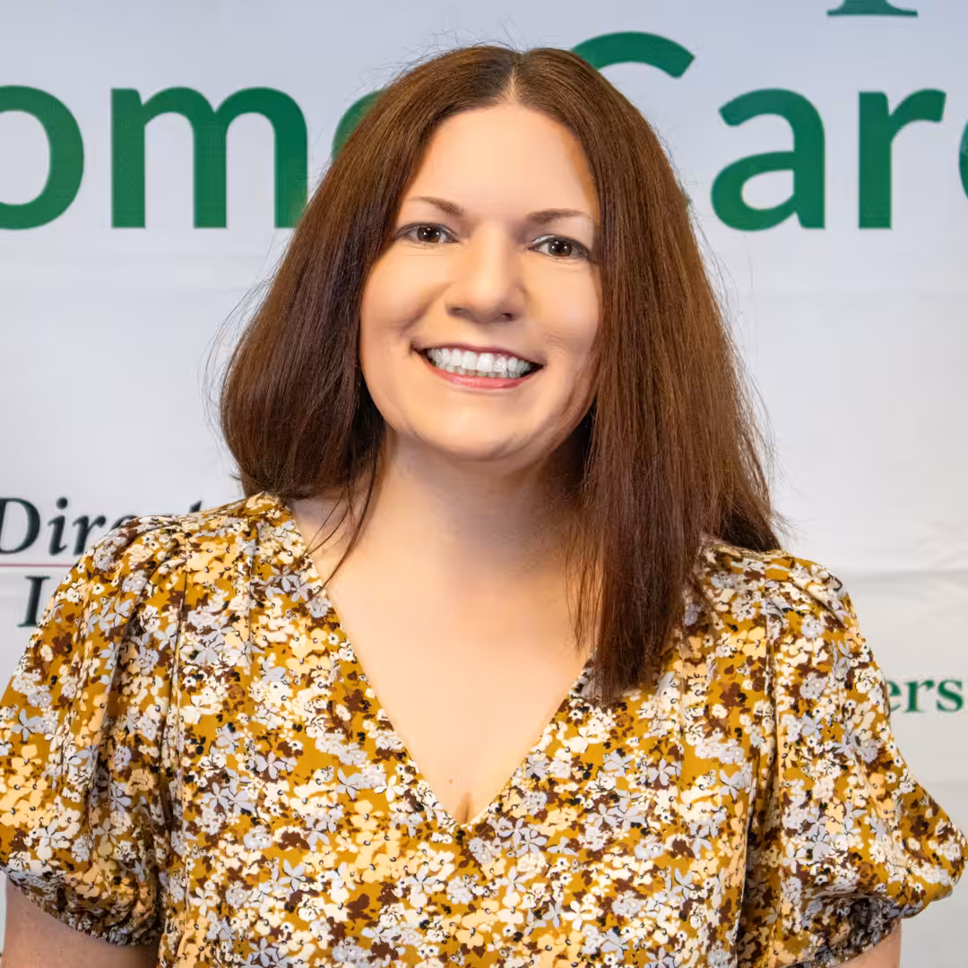 A woman with straight brown hair, wearing a yellow floral blouse, smiles at the camera in front of a banner with partial green text.