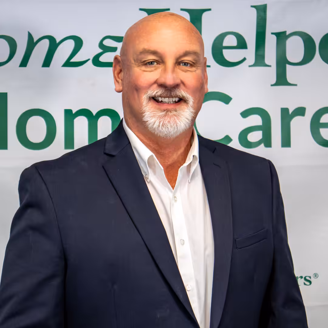 A smiling bald man with a white goatee is wearing a dark suit jacket and white shirt, standing in front of a banner with green text that partially reads Home Helpers Home Care.