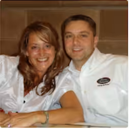A smiling woman and man sit close together, both wearing white shirts. The man has short hair and a name tag on his shirt. They are posed indoors in front of a brown wall.