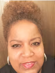 A woman with short curly hair, wearing hoop earrings and a black top, smiles at the camera in front of a wooden background.