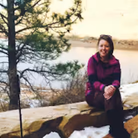 A woman in a purple jacket and patterned pants sits on a stone wall near a lake, with snow patches on the ground and a pine tree beside her. The background shows water and a sunlit hillside.