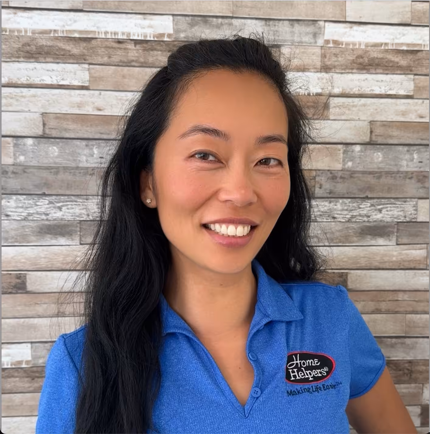 A woman with long black hair smiles at the camera. She is wearing a blue Home Helpers polo shirt and stands in front of a wall with a wood panel design.