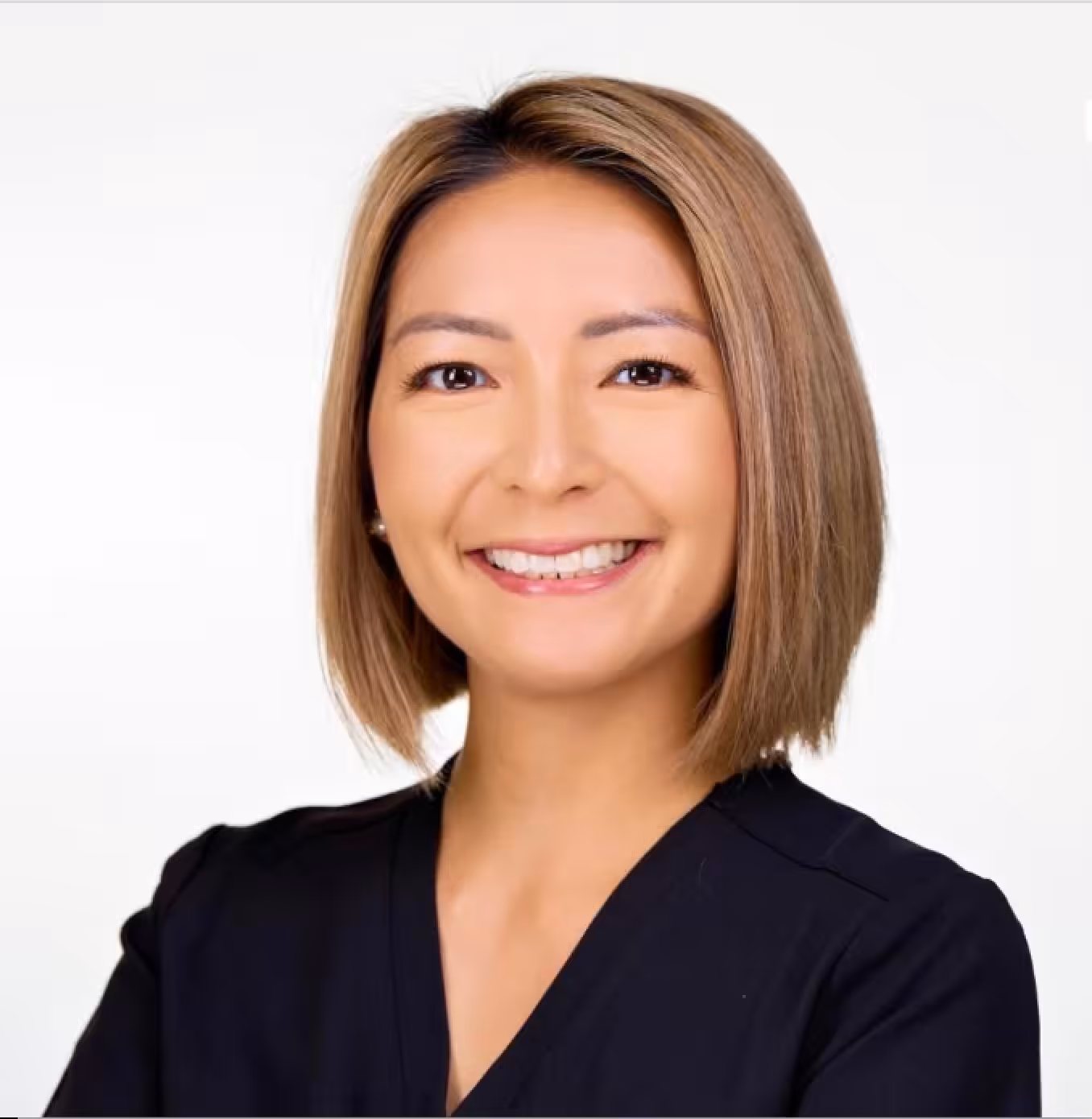 A woman with straight, shoulder-length light brown hair smiles at the camera. She is wearing a black top and stands against a plain white background.