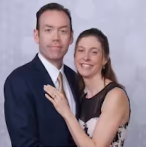 A man in a suit and a woman in a sleeveless dress stand close together, smiling, in front of a light-colored, patterned backdrop.
