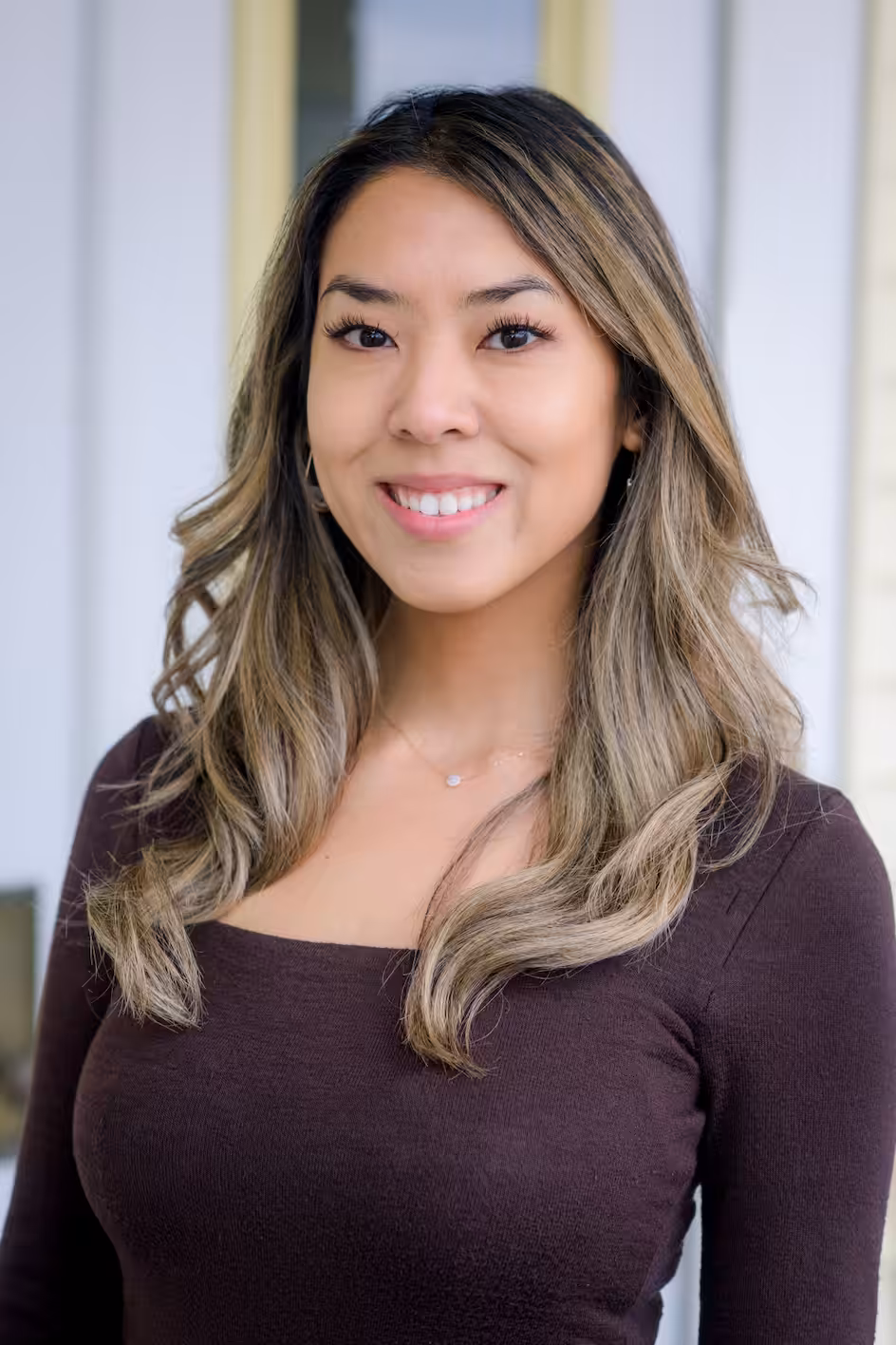 A young woman with long, wavy, light brown hair wearing a dark brown, long-sleeve top smiles at the camera while standing in front of a light-colored background.
