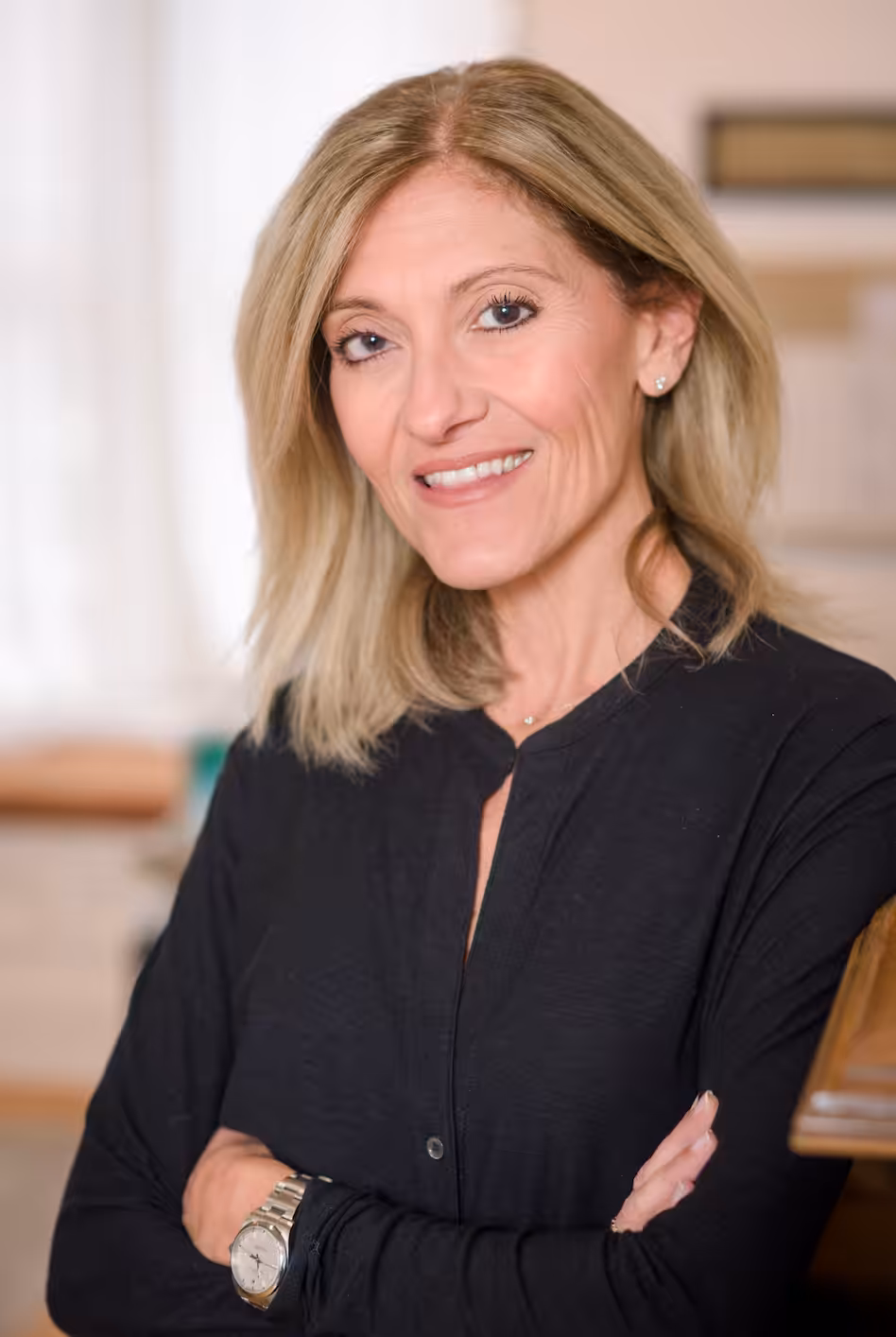 A woman with shoulder-length blonde hair, wearing a black blouse and a watch, stands indoors with her arms crossed, smiling at the camera. The background is softly lit and out of focus.