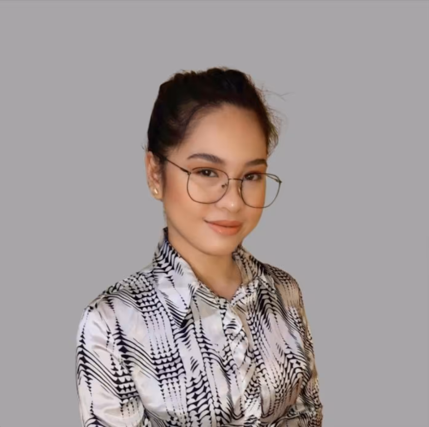 A woman with glasses and tied-back hair is smiling slightly. She is wearing a patterned blouse with black and white designs. The background is plain gray.