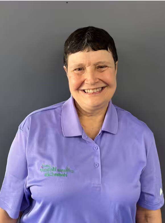 A person with short dark hair smiles at the camera, wearing a lavender polo shirt embroidered with the words Home Instead Home Care, standing against a plain gray background.