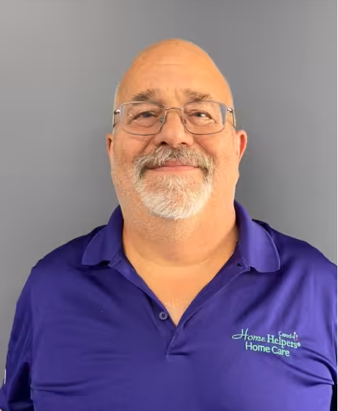 Smiling older man with glasses, a trimmed white beard, and a receding hairline, wearing a purple Home Helpers Home Care polo shirt, standing against a plain gray background.
