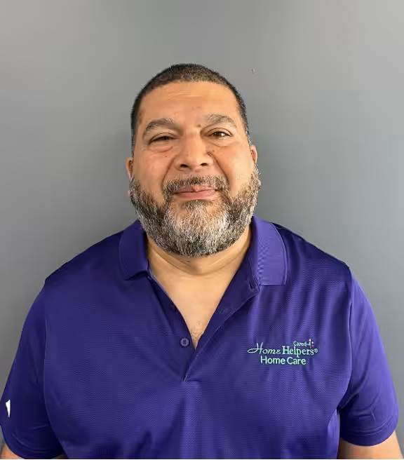 A man with short dark hair and a beard is smiling, standing against a gray background. He is wearing a purple polo shirt with the Home Helpers Home Care logo on it.