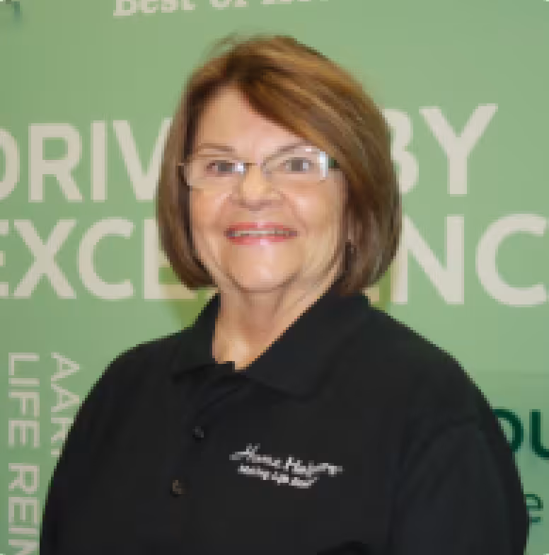 Smiling older woman with short brown hair and glasses, wearing a black collared shirt, standing in front of a green background with motivational text.