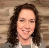 A woman with curly brown hair smiles at the camera. She is wearing a gray top and a light scarf, with a blurred wooden wall background.