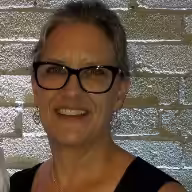 A woman with short gray hair and black glasses smiles in front of a silver brick wall. She is wearing a sleeveless black top and a delicate necklace.