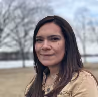 A woman with long brown hair and a beige jacket stands outdoors in a park with bare trees and a body of water in the background on a cloudy day.