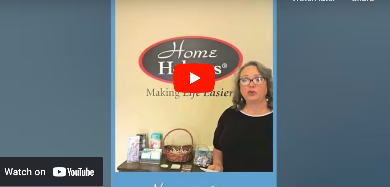 A woman stands beside a table displaying various items, in front of a yellow wall with a Home Helpers: Making Life Easier sign. A YouTube play button overlays the center of the image.