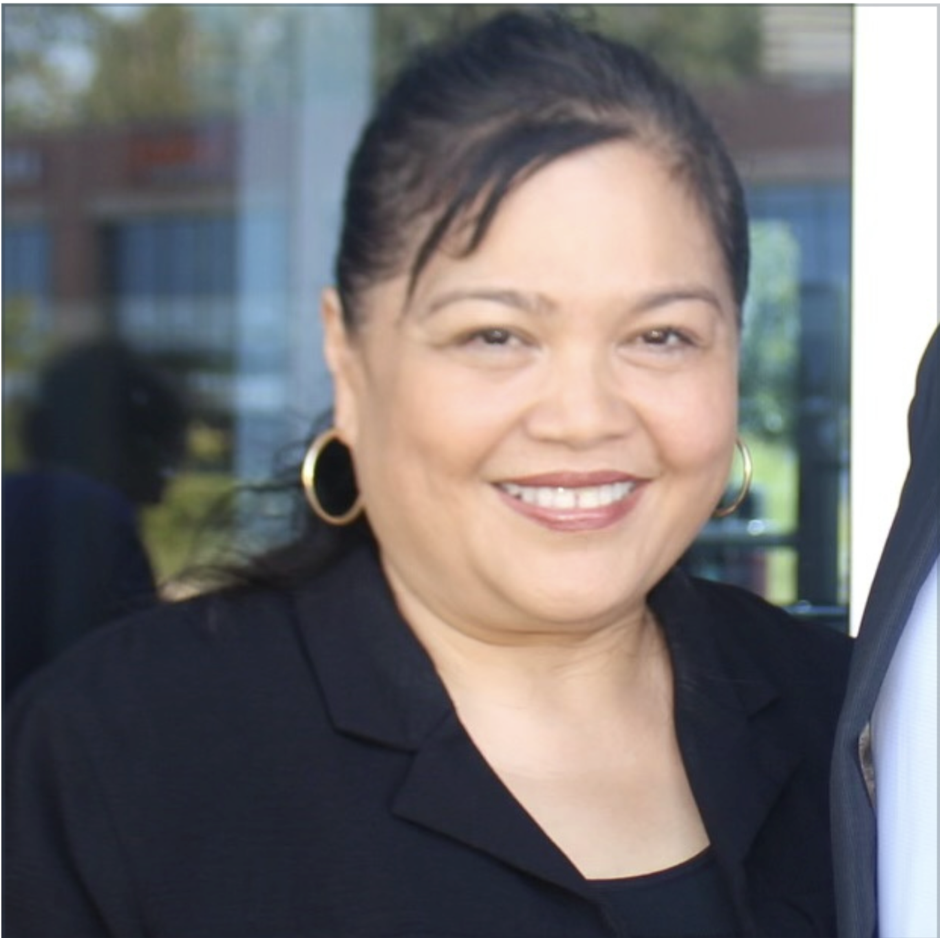 A woman with dark hair pulled back, wearing hoop earrings and a black top, smiles at the camera. The background shows glass windows and an outdoor setting.