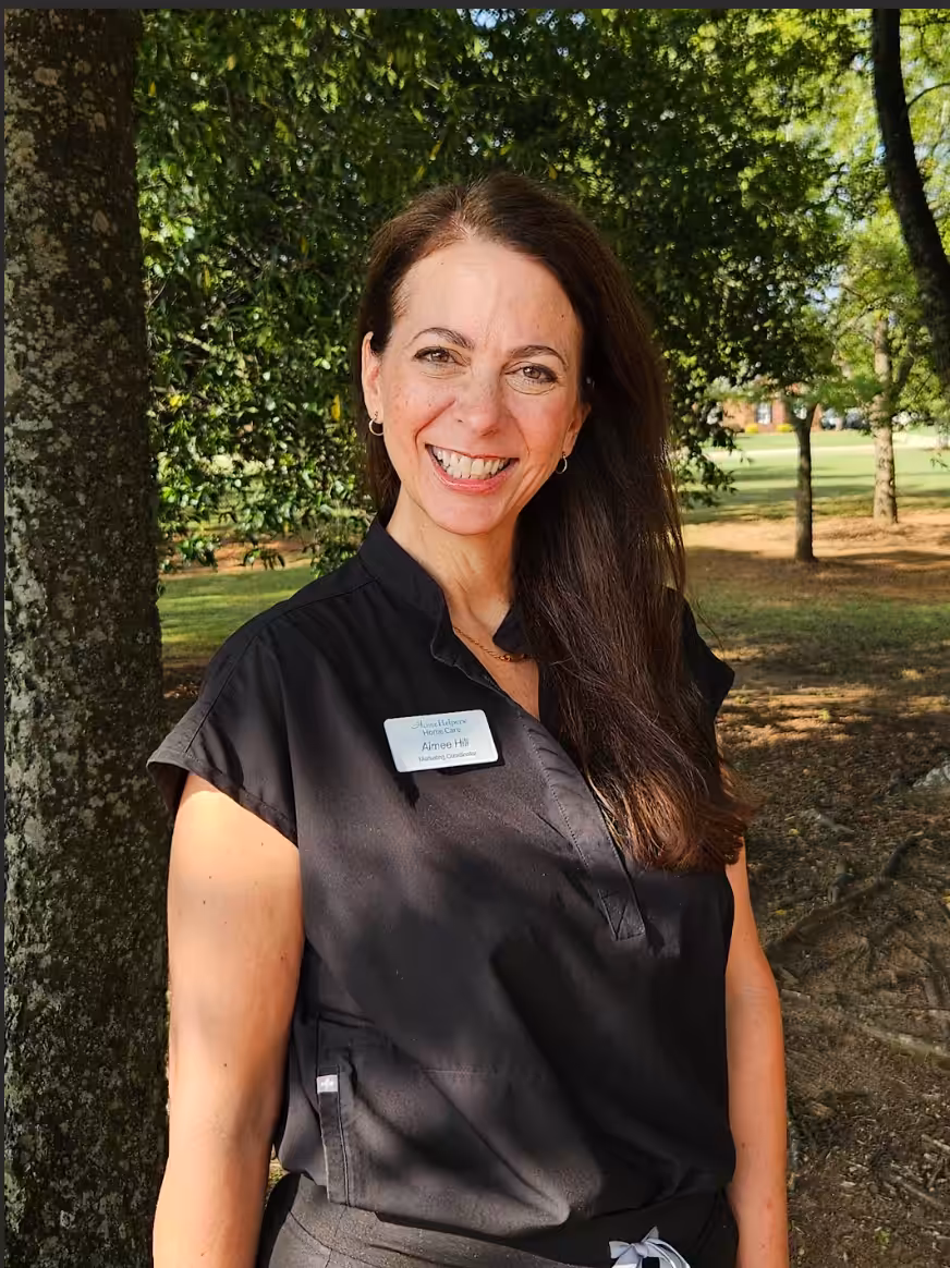 A woman with long brown hair, wearing a black short-sleeve blouse and a name tag, smiles while standing outdoors near a tree, with sunlight and greenery in the background.